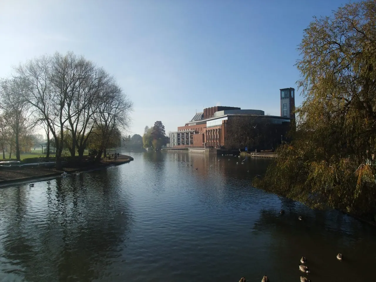 A brick theatre building with a tower stands beside a calm river lined with bare and autumn trees on a clear day.