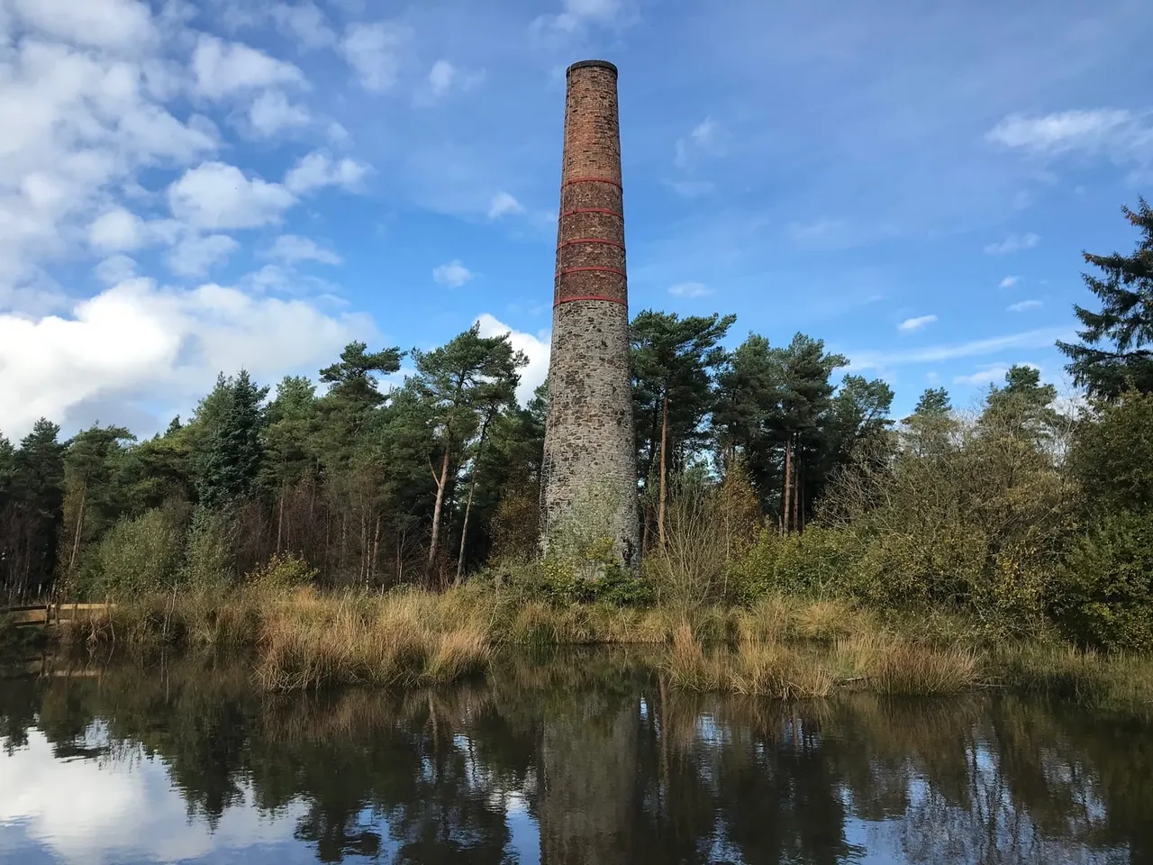 A tall brick chimney stands among trees beside still water under blue sky with white clouds.