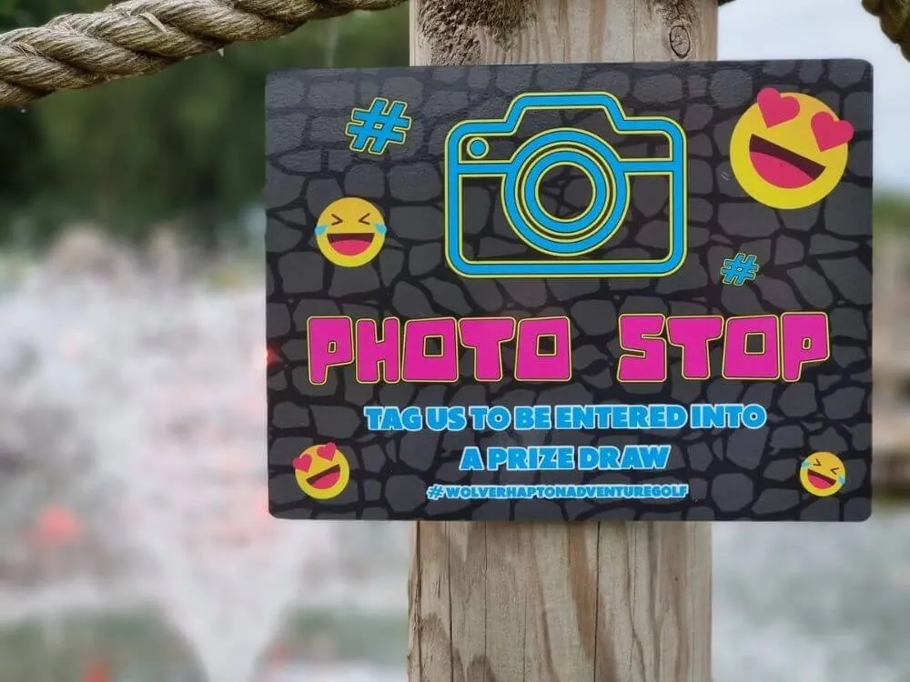 A colourful photo stop sign with neon graphics and emoji designs attached to a wooden post at a leisure venue.
