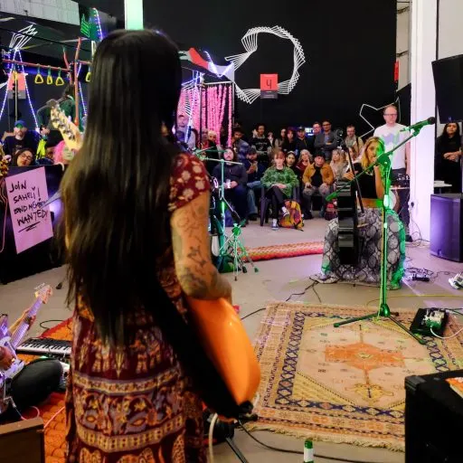 A band performs on stage with colourful neon decorations while a crowd watches from the venue floor.