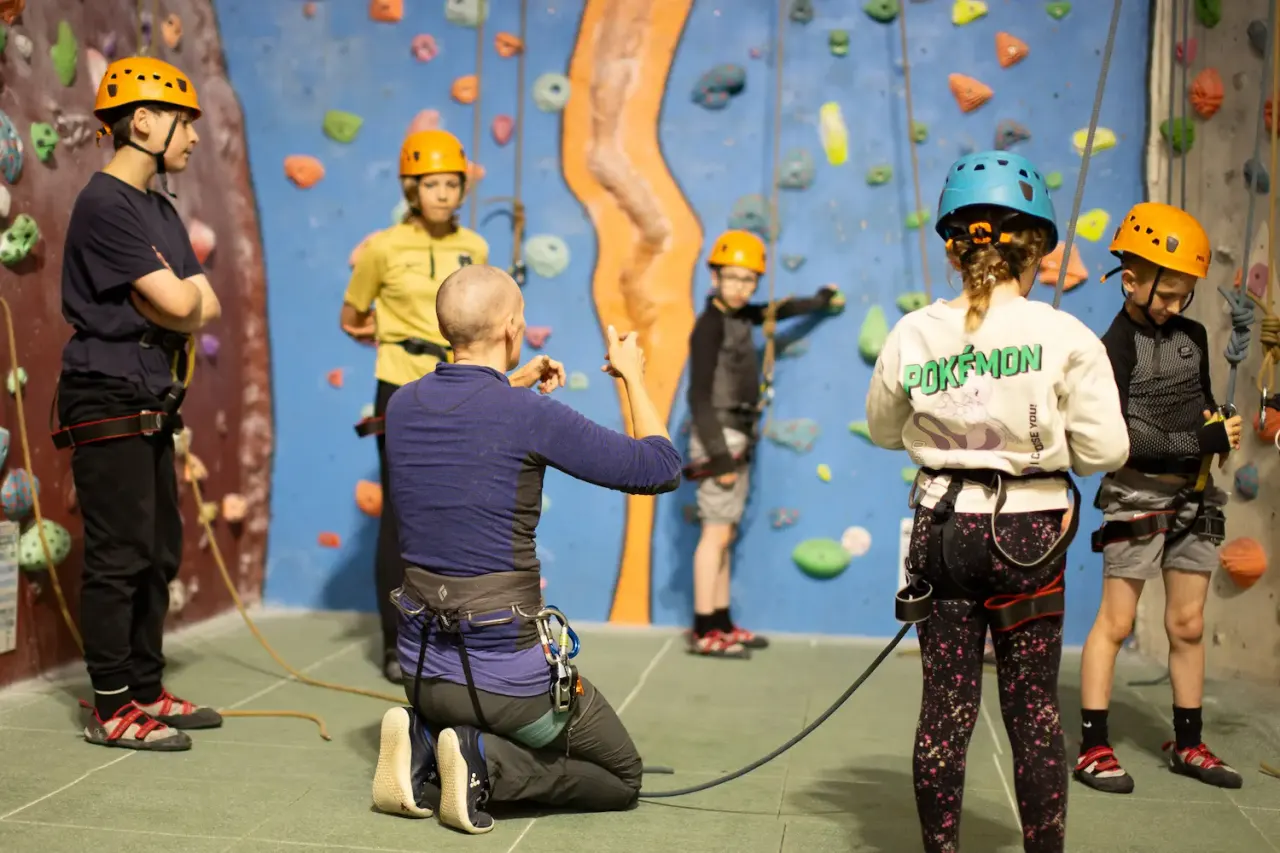 Multiple people of various ages wearing helmets whilst indoor rock climbing on a blue wall with coloured holds.