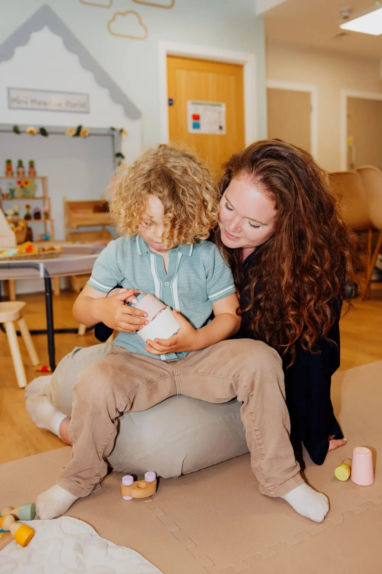 An adult and young child sit together on a mat in a bright playroom, looking at a small object the child is holding.