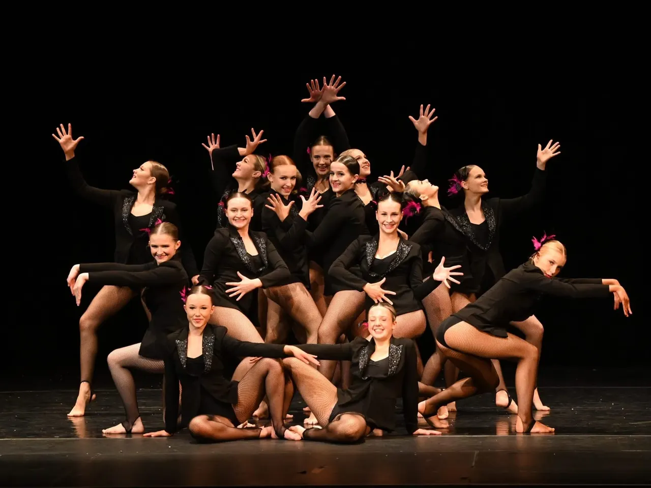 A group of young dancers in black outfits with pink hair accessories perform a choreographed routine on stage with raised arms and expressive poses.