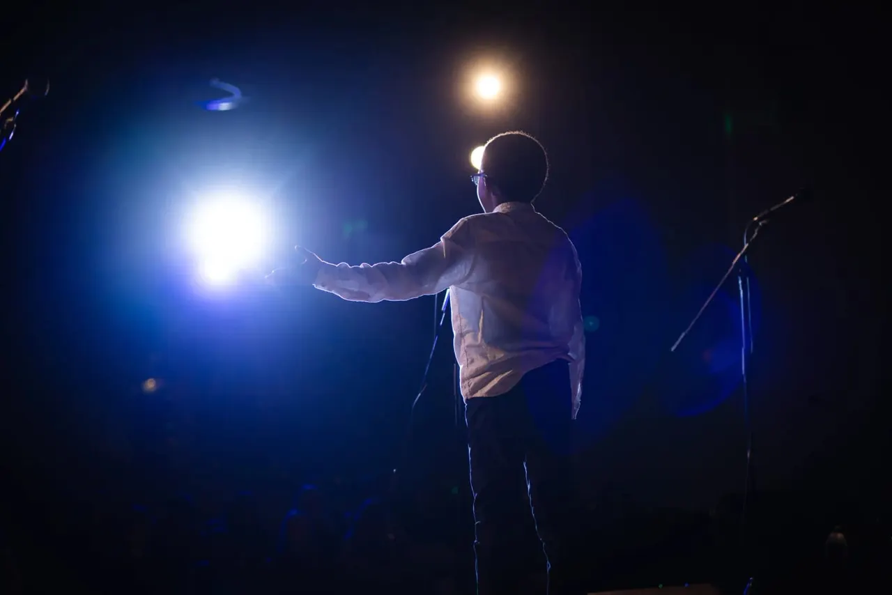 Performer on stage in spotlight, arms outstretched, with blue stage lighting and microphone stand visible.