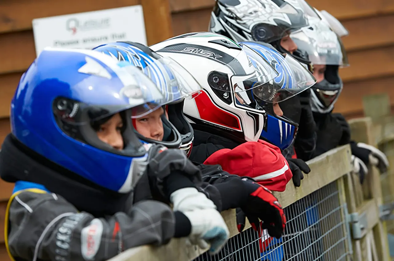 Kart racers in helmets and racing suits lined up at the barrier, ready to race.