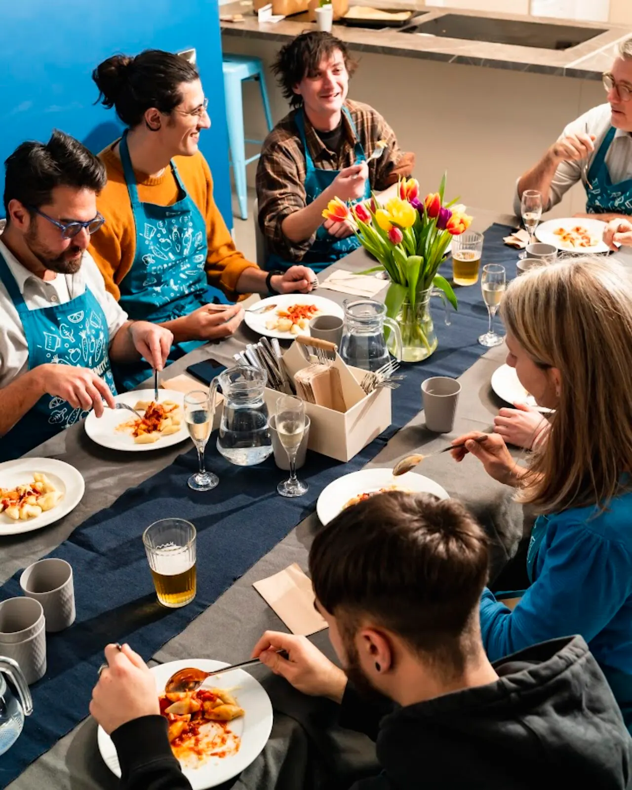 A group of people wearing blue aprons sit around a dining table eating a meal together, with staff serving and conversing with guests.