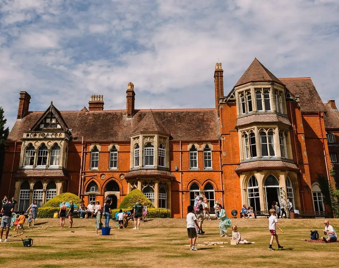 Visitors enjoying outdoor activities on the lawn in front of a Victorian red-brick mansion under blue sky.
