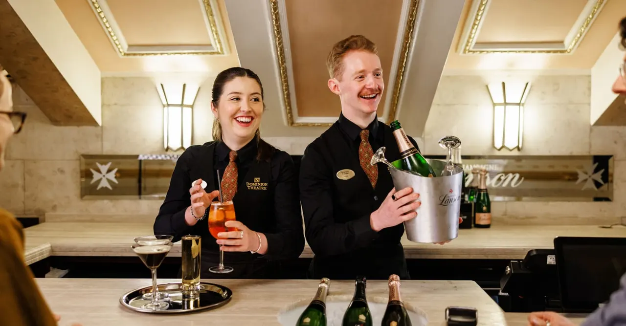 Two staff members smile behind an elegant bar counter, serving champagne and cocktails to guests in a ornate, gilt-decorated interior space.