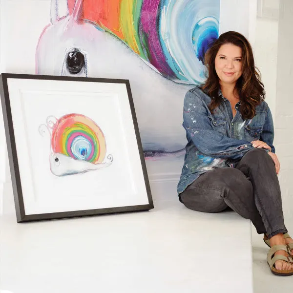 Woman posing next to colourful unicorn artwork and framed rainbow illustration in art studio.