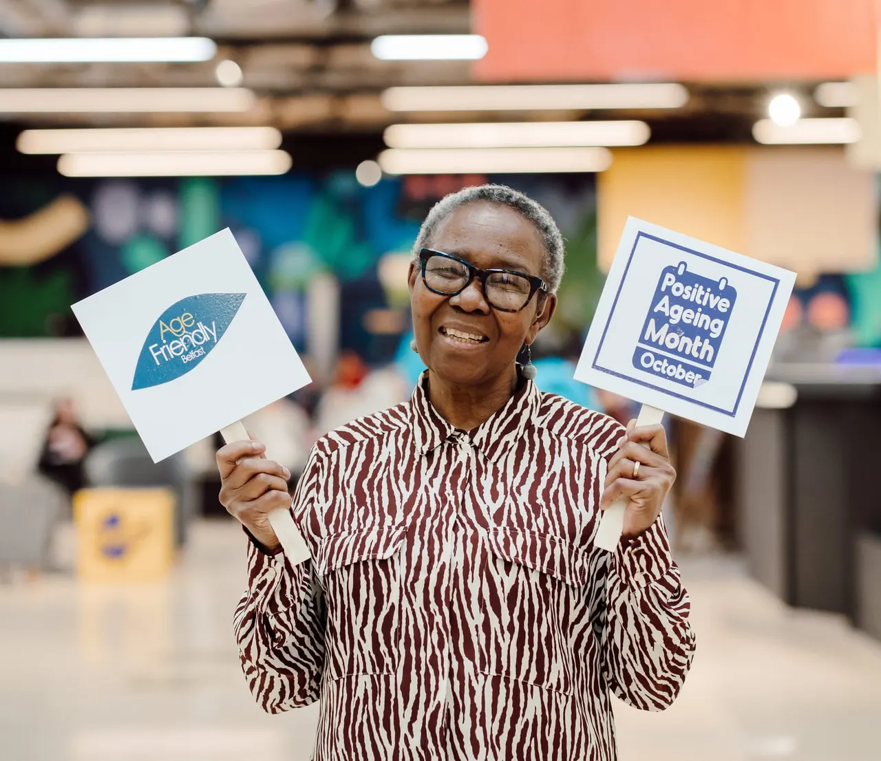 An older woman holding up Age Friendly and Positive Ageing Month signs, smiling at an indoor leisure venue.
