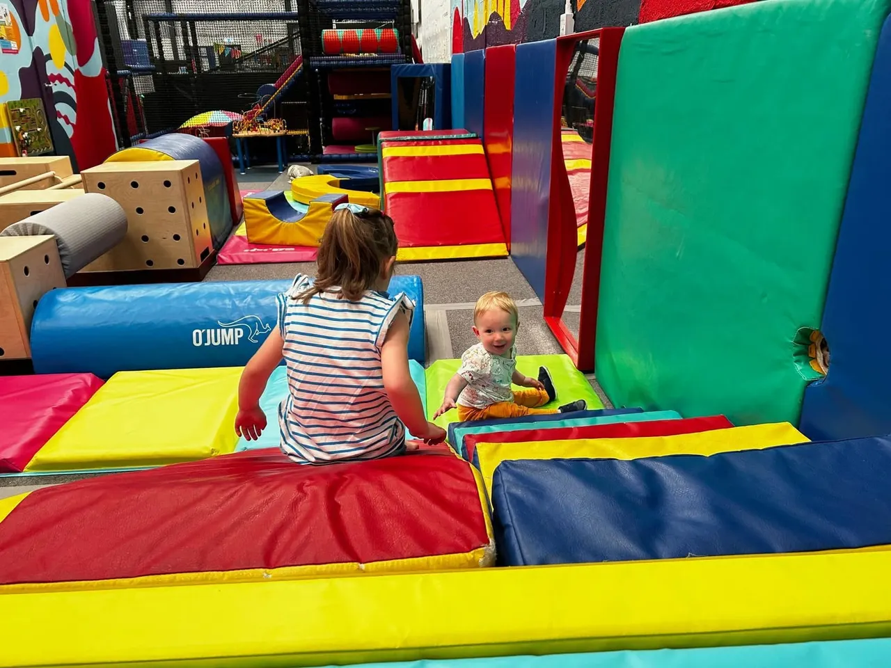 Two young children playing in a colourful indoor soft play area with padded equipment and obstacle courses.