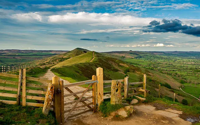 Wooden gate and fence in foreground overlooking a conical hill and patchwork farmland under a blue cloudy sky.