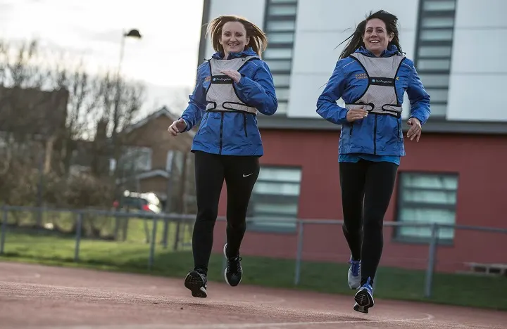 Two young runners in blue jackets jogging together on a sports track beside a leisure centre building.