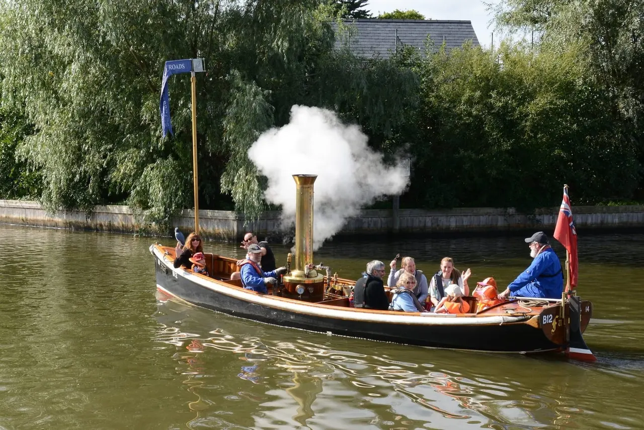 A traditional steam-powered narrowboat filled with passengers cruising along a tree-lined river, with white steam billowing from its chimney.
