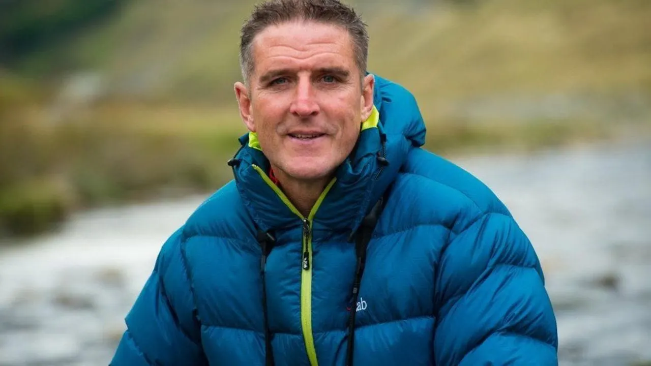 A man in a blue padded jacket smiles at the camera beside a river with blurred green landscape behind.