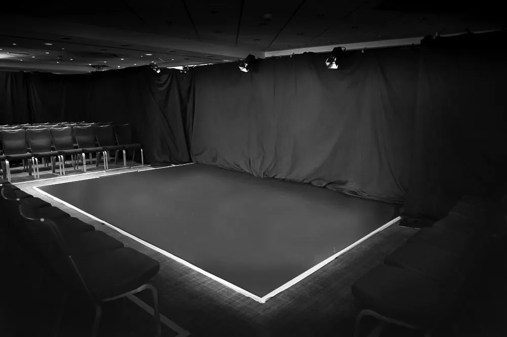 Empty black-walled performance or event space with lit stage platform and seating area.
