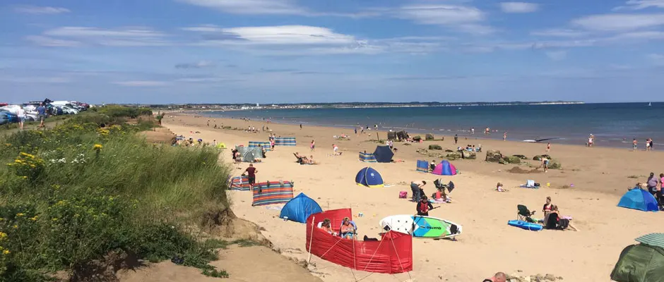 Busy sandy beach with dozens of visitors, colourful windbreaks and tents, swimmers in the sea, and grassy dunes along the shoreline.