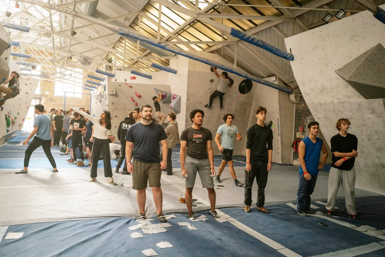 A group of people of mixed ages stand and wait inside an indoor climbing gym with high walls featuring coloured holds and blue padded beams overhead.