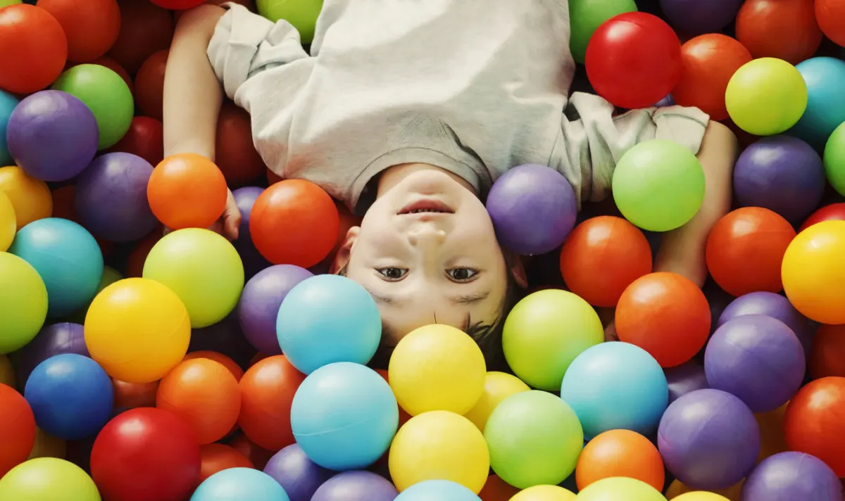 Child lying in a ball pit surrounded by colourful plastic balls, looking up at the camera.
