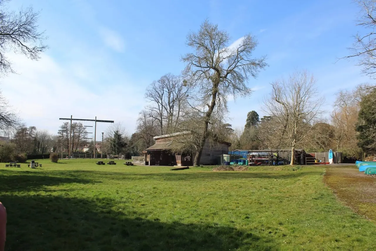 Open grass field with wooden buildings, picnic tables, play equipment and bare trees under blue sky.
