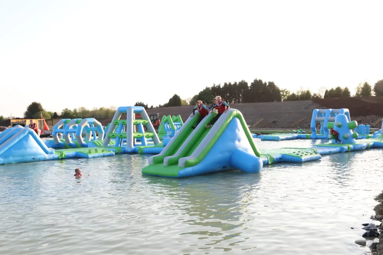 Children and adults enjoy inflatable water slides and obstacles in a lake surrounded by trees.