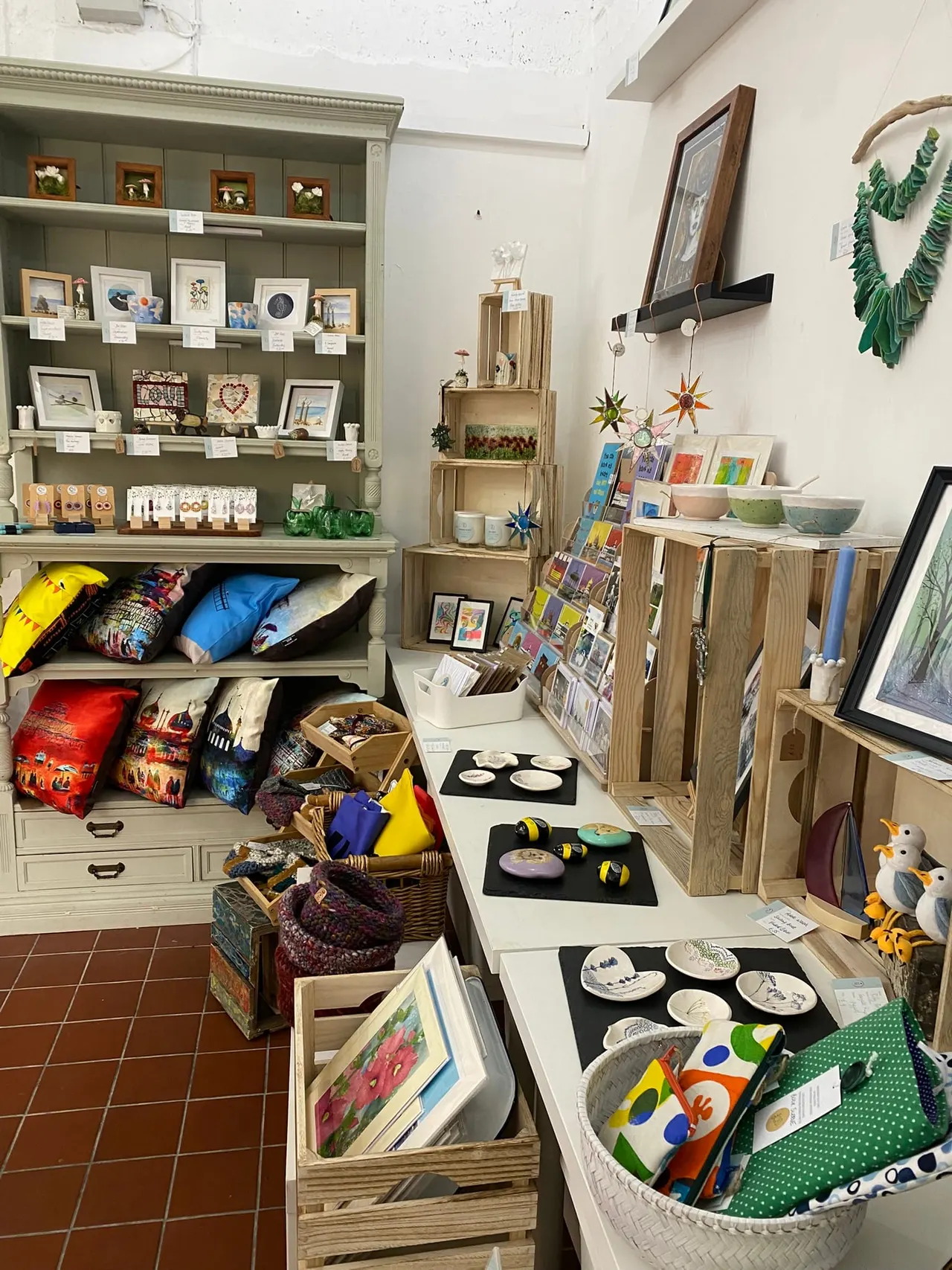 Interior of craft shop displaying shelving units with framed artwork, handmade ceramics, gift items, and colourful products for sale.