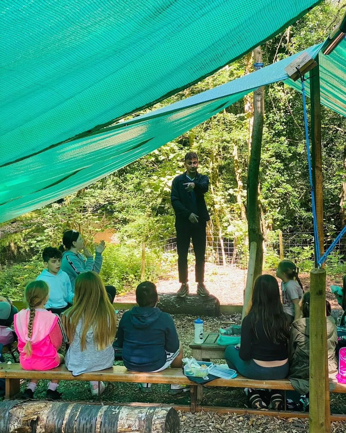 An adult instructor stands on a log addressing a group of children seated on a bench beneath a green shade sail in a woodland outdoor classroom setting.