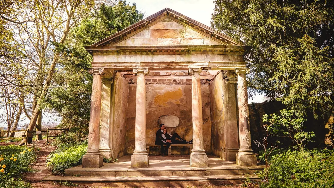 Two people sit on a bench inside a classical stone temple pavilion surrounded by woodland and gardens.