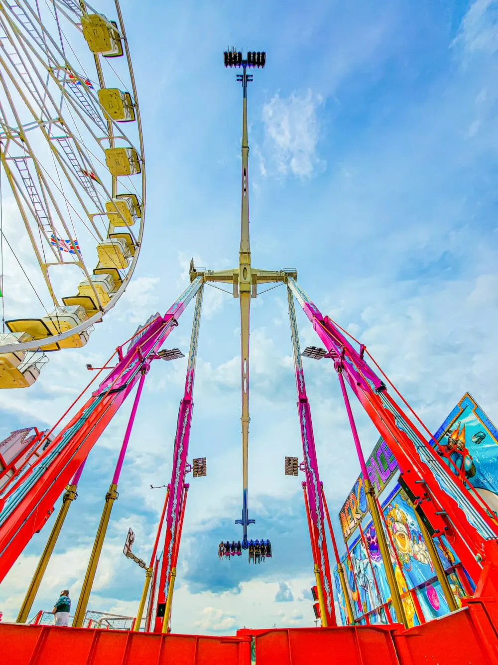 Upward view of fairground rides including a yellow Ferris wheel and pink and red swinging ship ride against blue sky.
