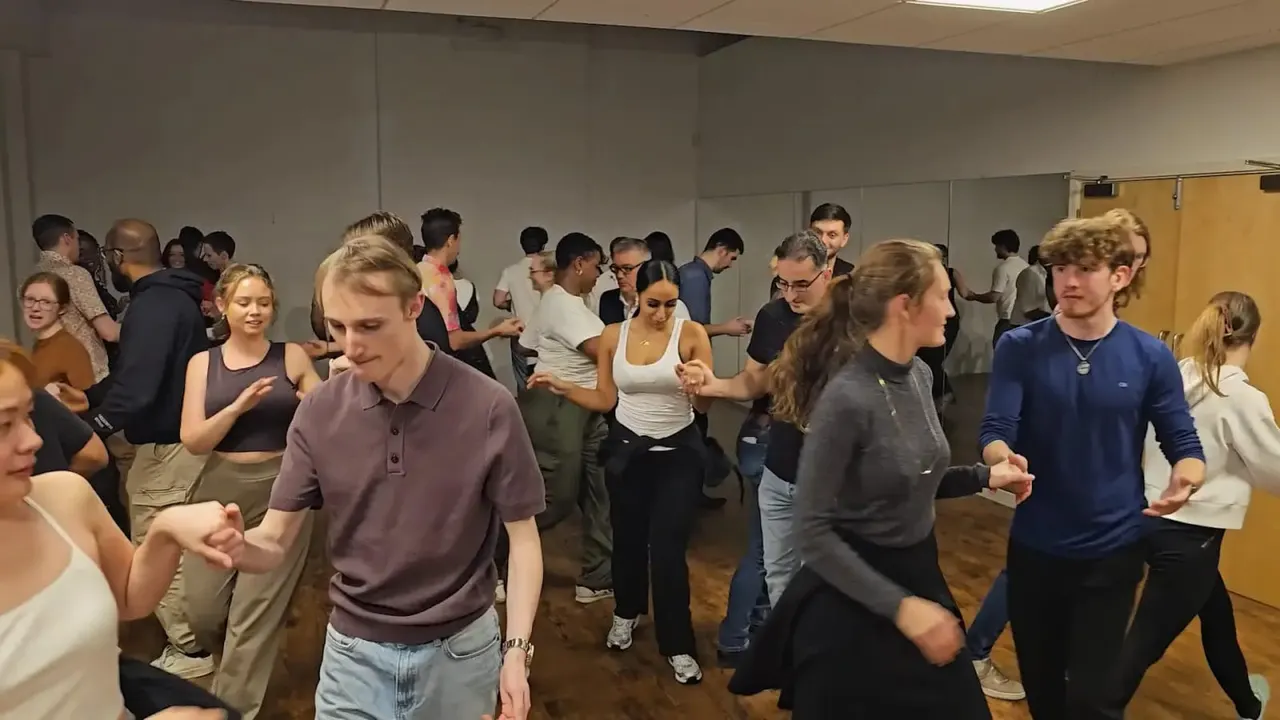 A large group of people dancing together in an indoor studio with wooden flooring.
