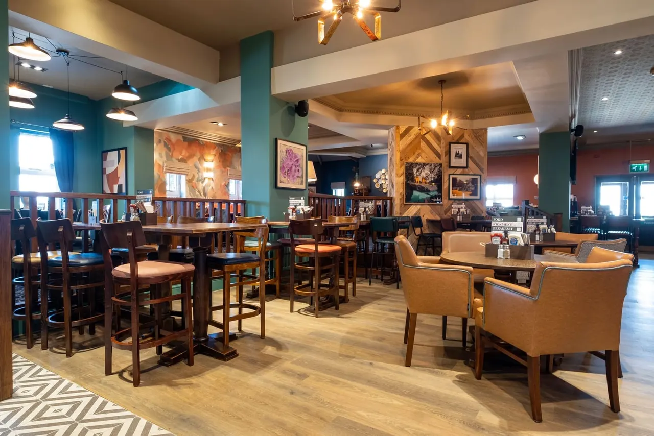 Modern hotel lounge with bar seating, high tables with chairs, and comfortable armchairs arranged throughout the space with teal and warm-toned décor.