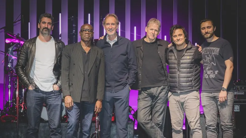 Six men stand together on stage with purple lighting and a drum kit in the background.
