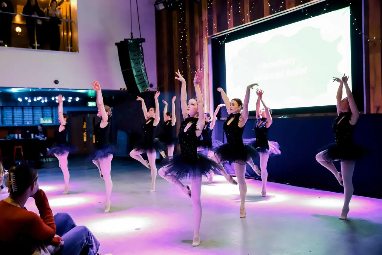 Young ballet dancers performing on stage in black outfits under coloured lighting in front of an audience.