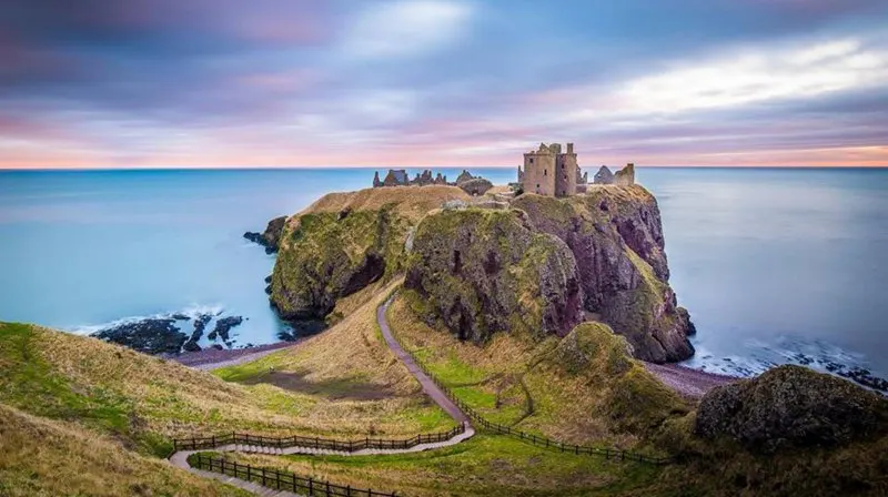 Ruined castle perched on a rocky island surrounded by sea, with a winding causeway and grassy slopes at dusk.