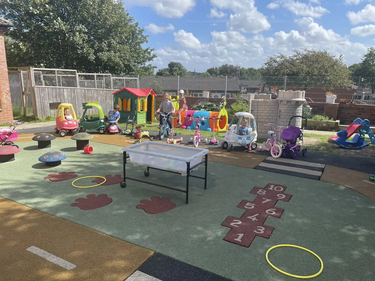 Young children playing in an outdoor nursery area with toy vehicles, a playhouse, and learning games on a sunny day.