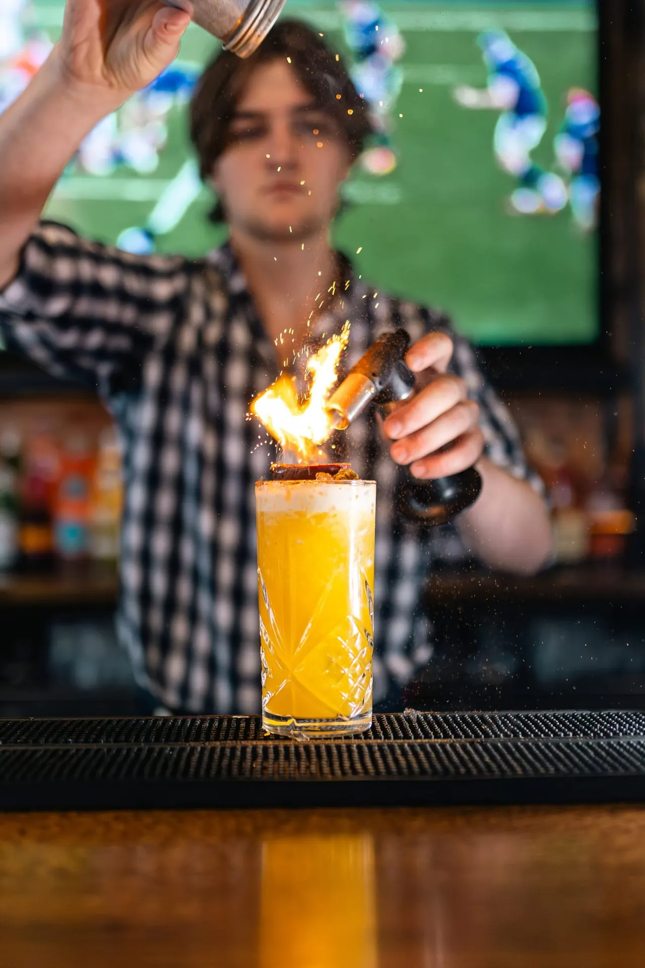 Bartender igniting a flaming cocktail at a bar counter with a sports screen visible in the background.