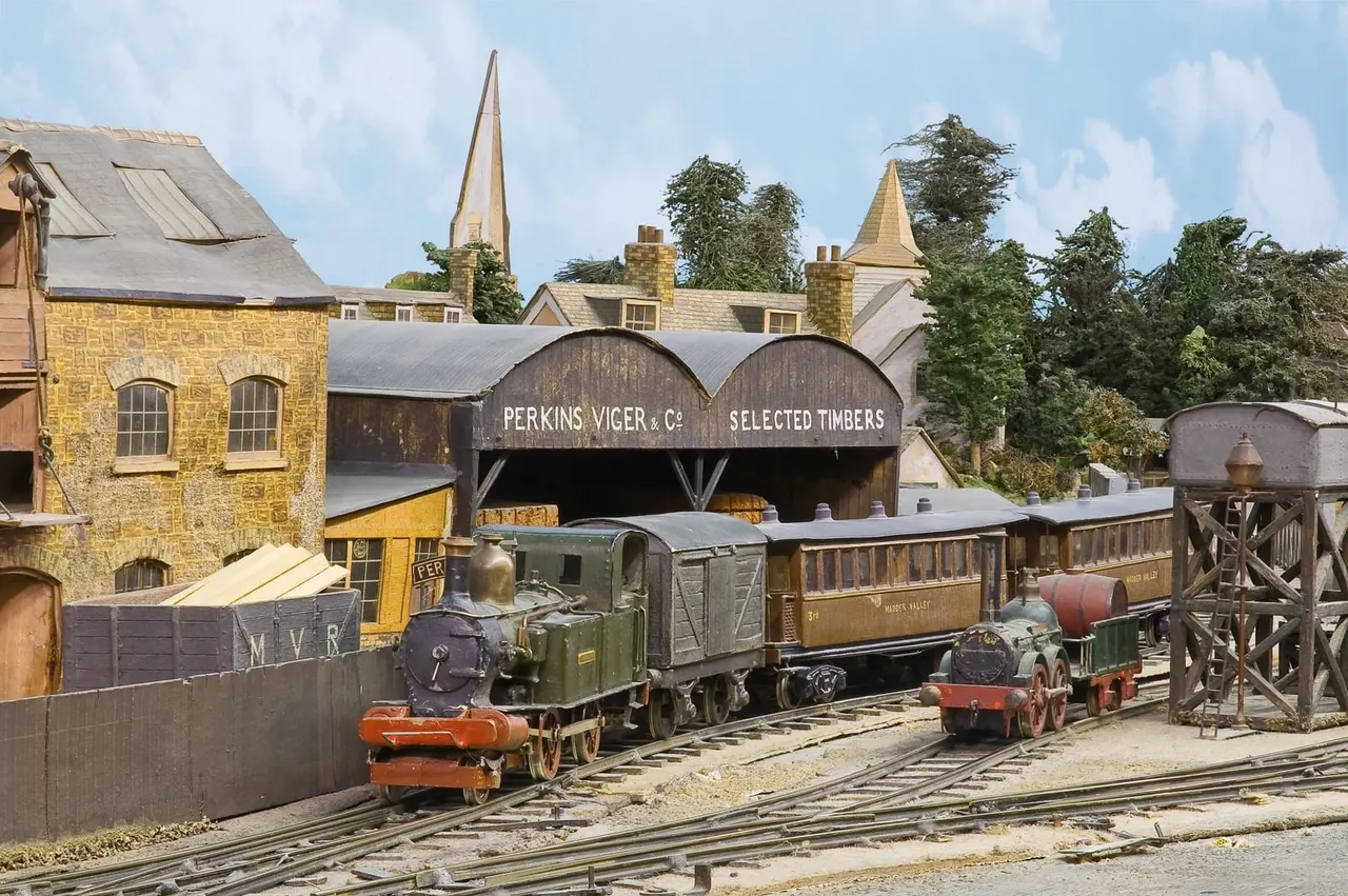 A vintage narrow-gauge steam locomotive with passenger and goods carriages at a heritage railway station with period buildings and timber yard.