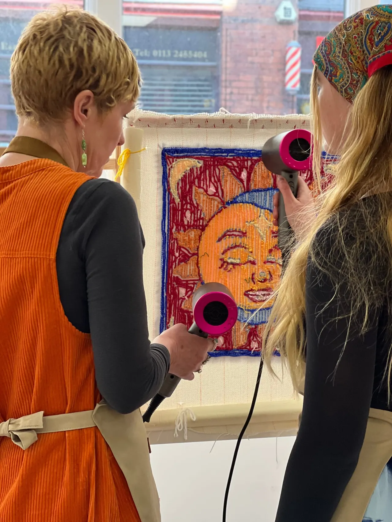 Two people using pink handheld tufting guns to create a colourful textile artwork on a wall-mounted canvas frame.