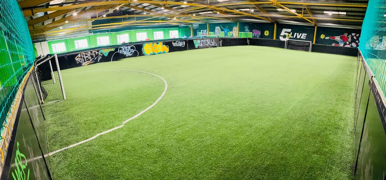 Empty indoor five-a-side football pitch with bright green artificial turf, surrounded by netting and advertising boards.