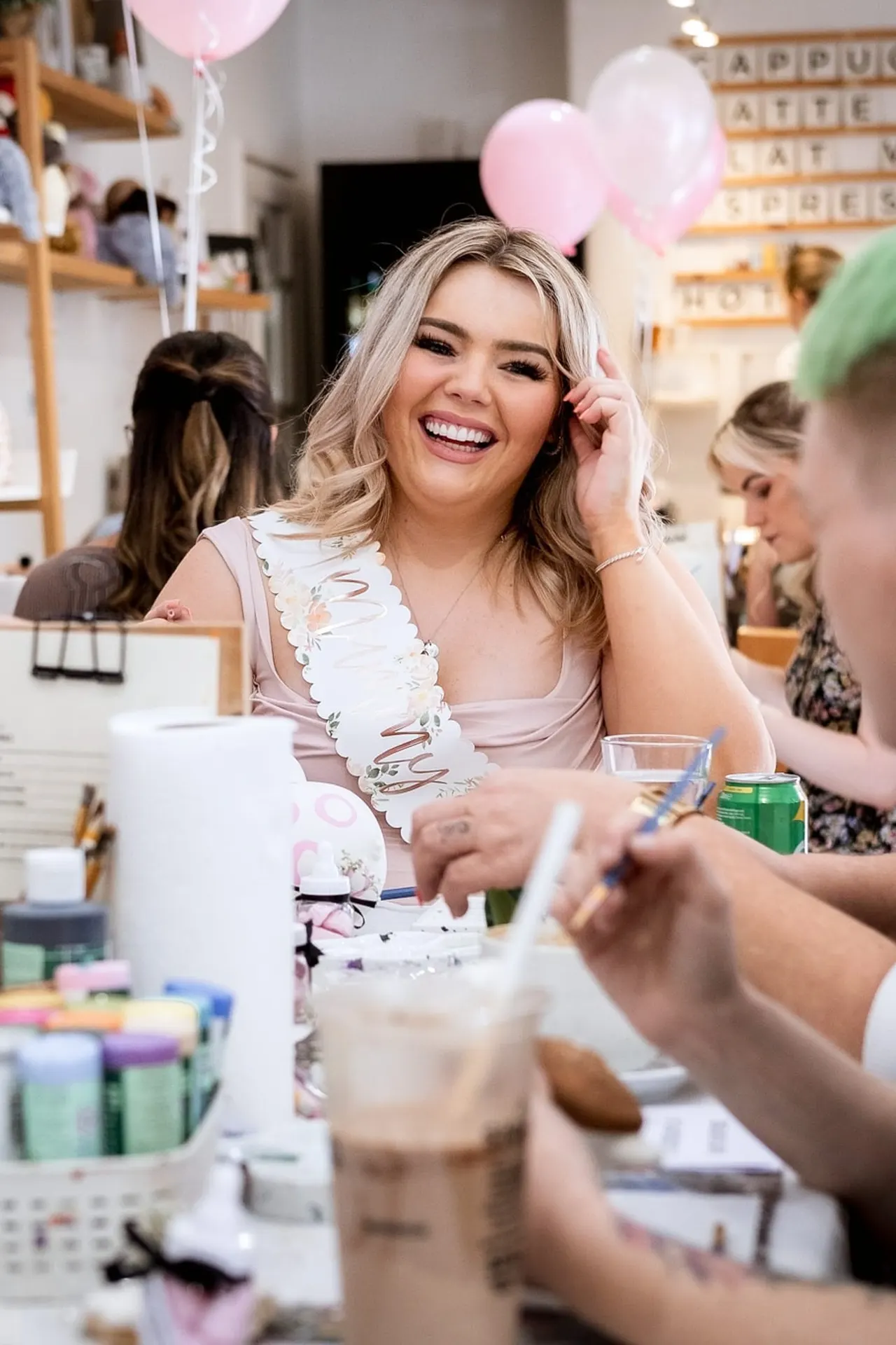 Young woman smiling at a craft table surrounded by paints, brushes and pottery supplies, with pink and white balloons overhead.