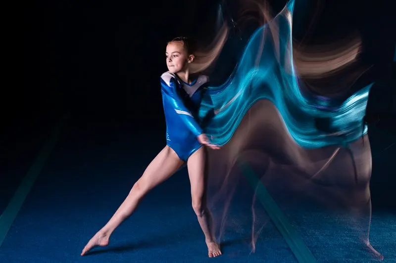 Young gymnast performing rhythmic gymnastics with flowing blue ribbon against dark background.