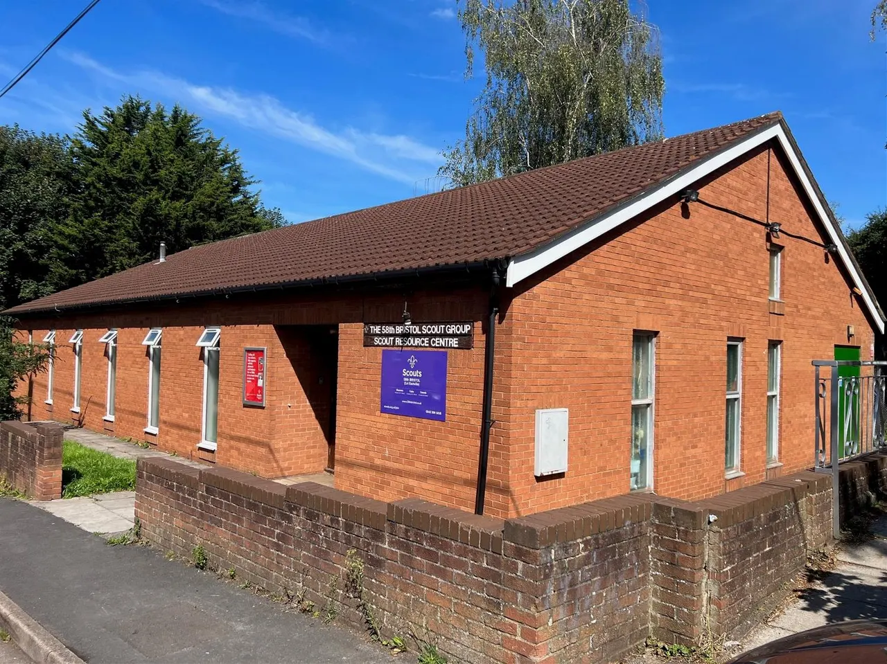 Single-storey red brick scout building with brown tiled roof, surrounded by green trees under blue sky.