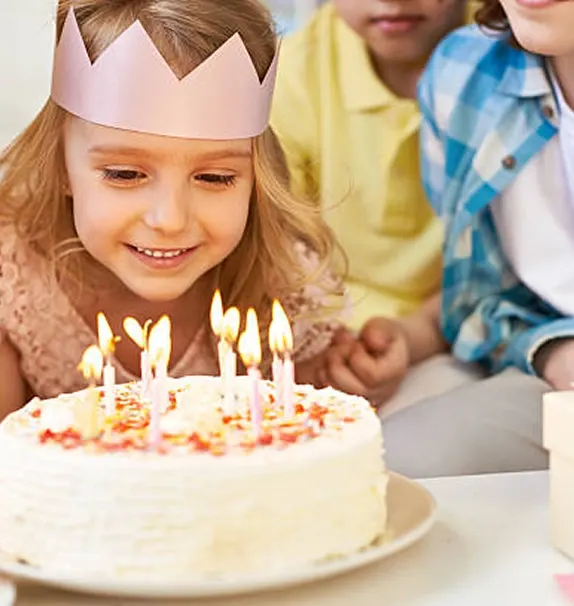 Young child wearing paper crown blows out candles on birthday cake whilst other children watch.