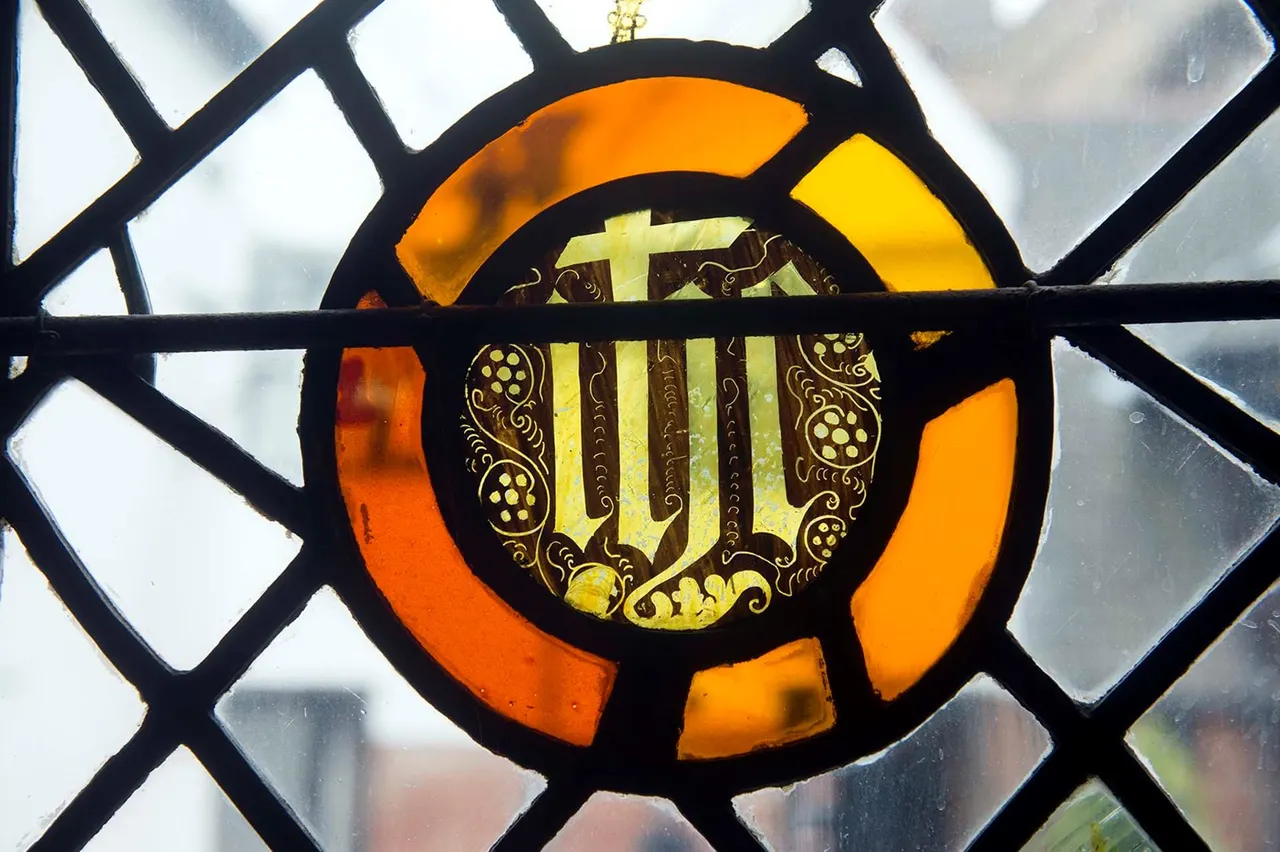 Stained glass window detail showing a decorative circular medallion with a yellow cross and ornate gold scrollwork against orange and clear glass panes.