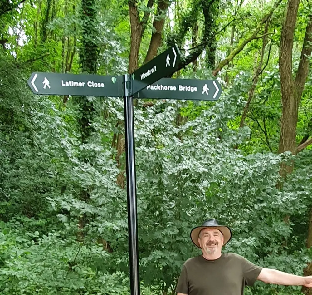 Man standing beneath woodland directional signpost with arms outstretched, surrounded by dense green foliage and tall trees.