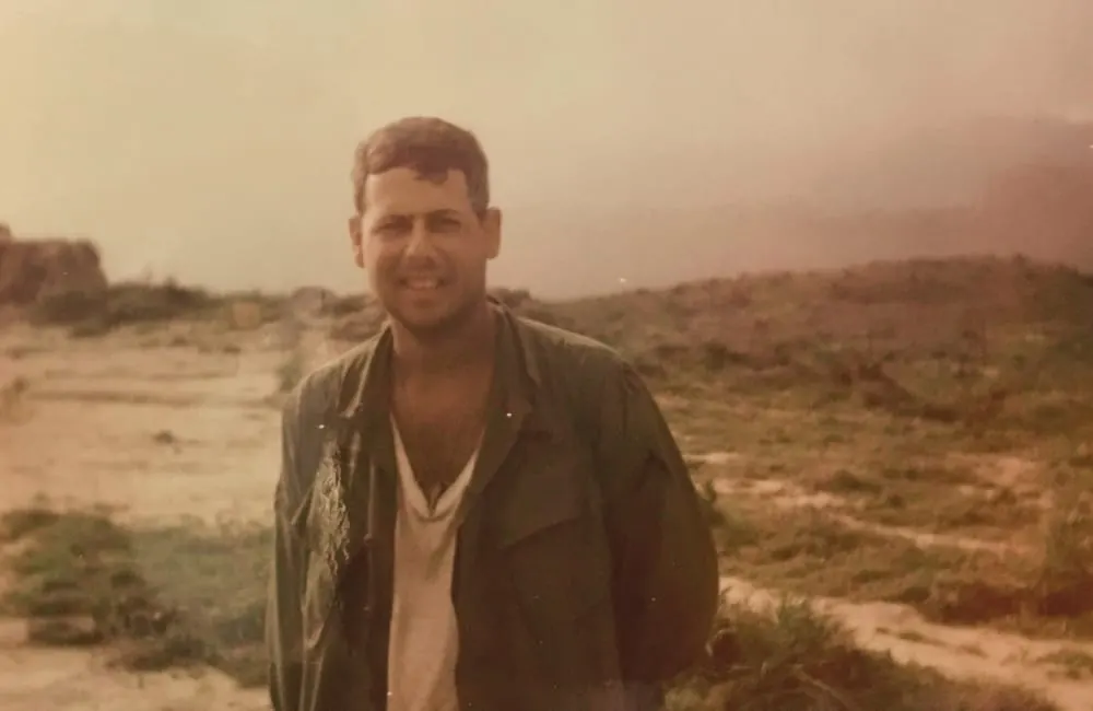 A man smiling at the camera in an arid, sparsely vegetated desert landscape at dusk.