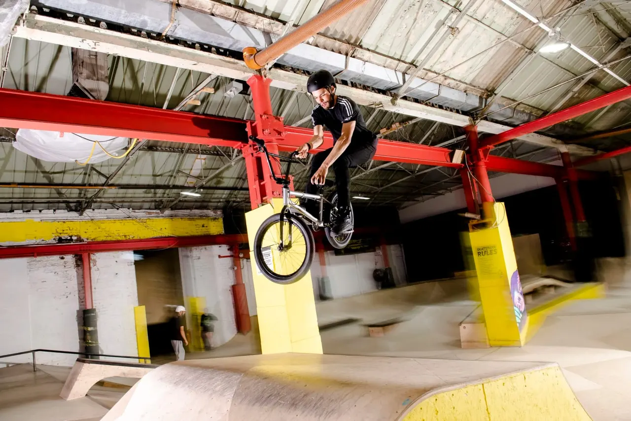 Person performing a jump on a BMX bike over a yellow ramp in an indoor skate park with red steel beams.
