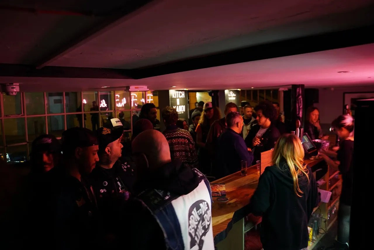 Crowd of people socialising in a dimly lit bar with pink-red lighting and neon signs.
