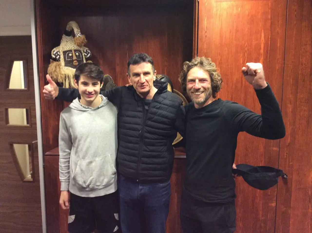 Three visitors pose cheerfully indoors next to a large decorative bird figure mounted on a wooden wall.
