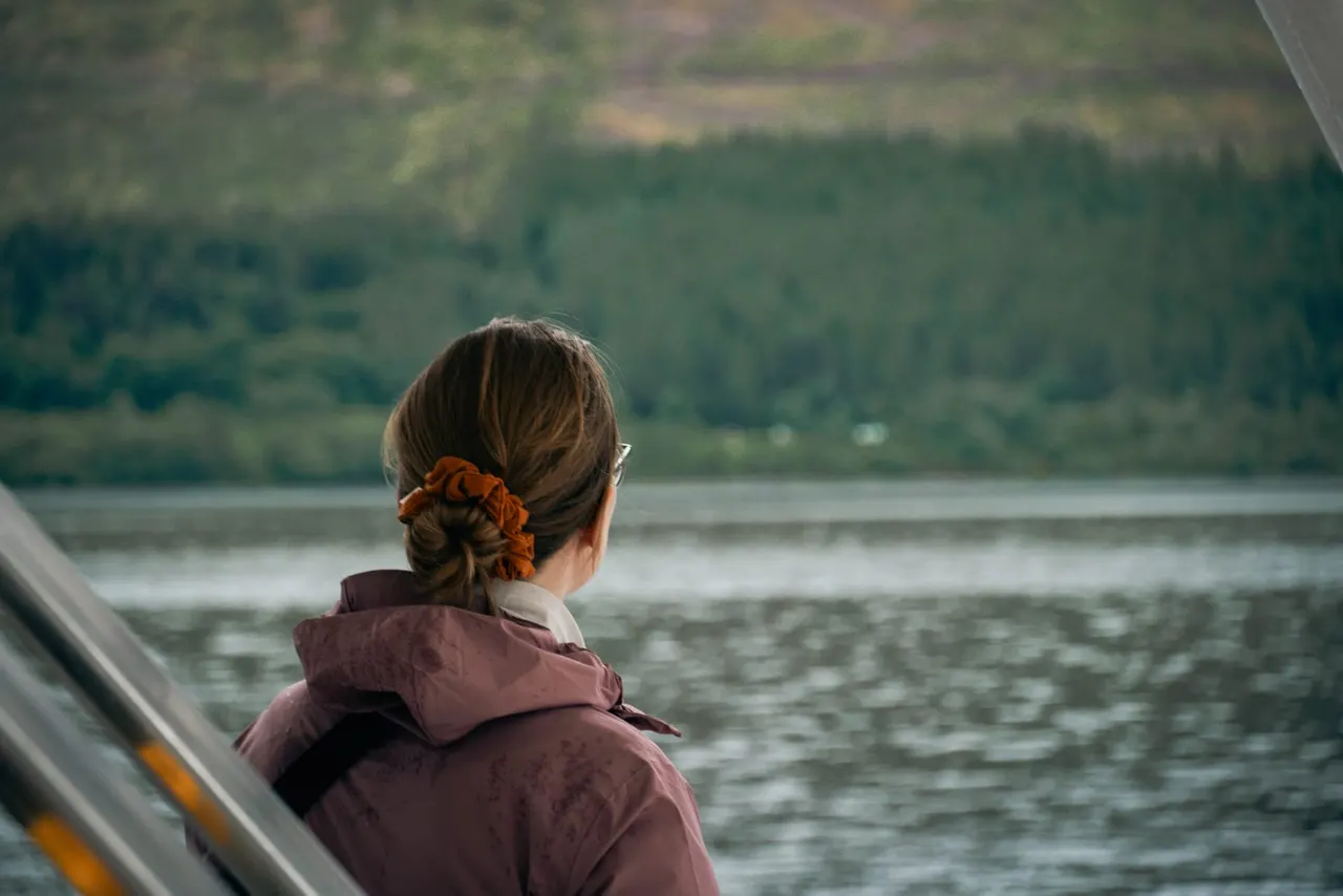 Person viewing a loch from a boat, with forested hills in the background.